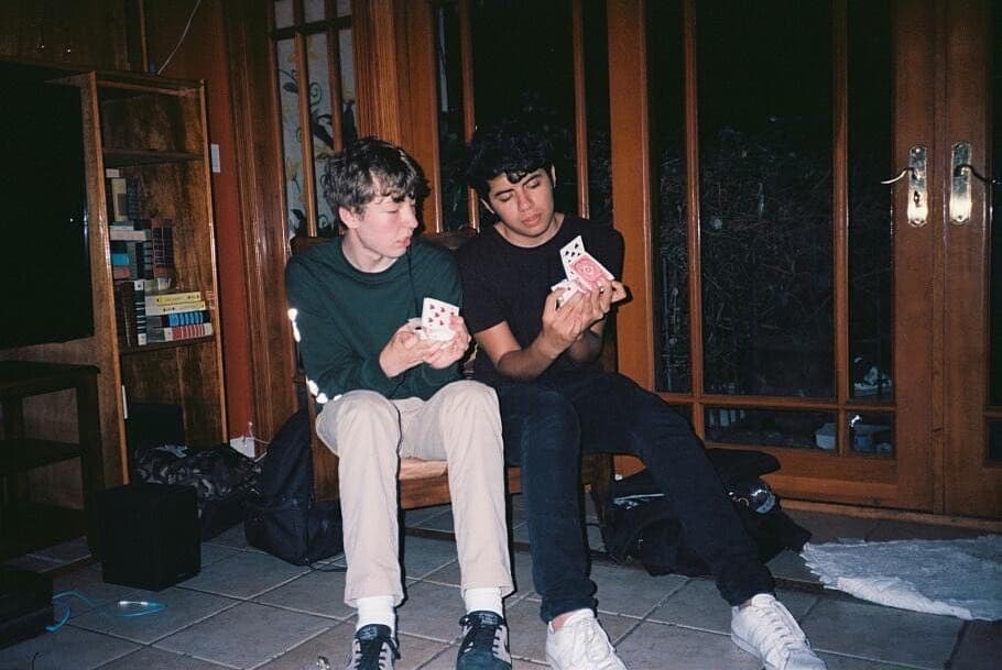Two cardists sitting side by side on a bench at night, both mid-flourish, string lights behind them.